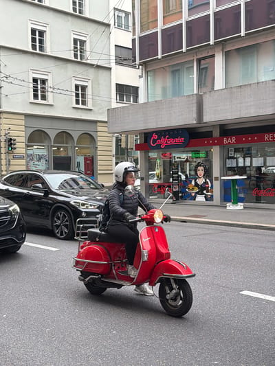 A person is riding a red Vespa scooter on Stadthofstrasse in Lucerne, Switzerland. The scooter is positioned near the curb in front of a black car. Behind the scooter and car is the "California Bar Restaurant" building with signage including logos for Carlsberg and Coca-Cola. A hand-drawn advertisement depicting a woman holding food is affixed to the glass door. The restaurant's sign reads "California". The building across the street has several windows. Electrical lines run along the side of the building on the left. A set of traffic lights is visible.