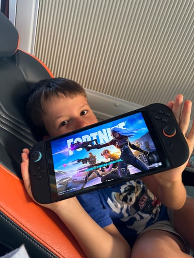 A young boy is lying on a black and orange chair while holding a handheld gaming device. The screen displays the game "Fortnite." The boy is looking at the screen with visible interest. The background consists of a window covered with blinds. The location is inside a room, but there is no specific indication of the city of Phan Son, Vietnam.