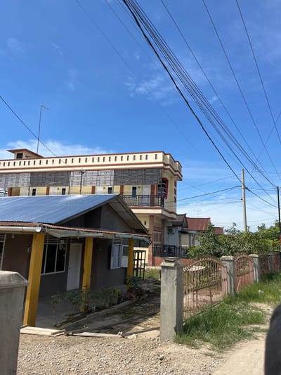 The image shows a street scene in Dalam, Indonesia. In the foreground, there is a gravel road and a low fence with decorative metal bars. Several houses and buildings are visible, varying in size and architectural style. One building, prominently featured on the left, has a dark gray facade with yellow pillars and a corrugated metal roof. To its right, a larger, multi-story building with a yellow and cream-colored exterior features decorative latticework on its windows and a cream-colored cornice. Further down the street, other buildings with red roofs and similar residential structures are visible. Overhead, a network of power lines and an antenna extend across the bright blue sky, which is dotted with a few white clouds. The overall scene suggests a residential or mixed-use urban environment with ongoing construction or development, indicated by the gravel road and visible building materials.