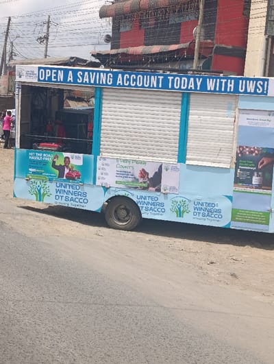A mobile financial service unit, colored light blue and white, is parked on the unpaved shoulder of an asphalt road in Matungulu west ward, Kenya. The unit, possibly a modified truck or trailer, displays prominent signage reading "OPEN A SAVING ACCOUNT TODAY WITH UWS!" and "UNITED WINNERS DT SACCO Growing Together" along its sides. Various advertisements promoting savings and wealth growth are also visible. The unit features an open section on the left, revealing interior equipment, and a closed white roll-down shutter in its central portion. A single black wheel is visible on the vehicle's right side. A person in a pink shirt stands to the left of the open section of the unit, facing inwards. In the background, a multi-story building with red and black patterned roofing and siding is prominent, surrounded by utility poles with an intricate web of overhead wires. The sky appears overcast.