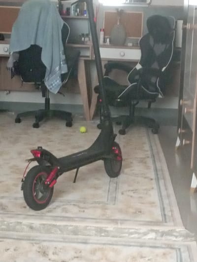 An electric scooter is parked on a rug in a room. The scooter is black with red accents on the wheels and taillight. Behind the scooter, there are two office chairs. One chair is covered with a light blue blanket. On a shelf in the background, there are several bottles. A small yellow ball is on the rug near the scooter. The scene appears to be an indoor residential or office setting. The image does not contain any information to determine its location in Keles, Uzbekistan.