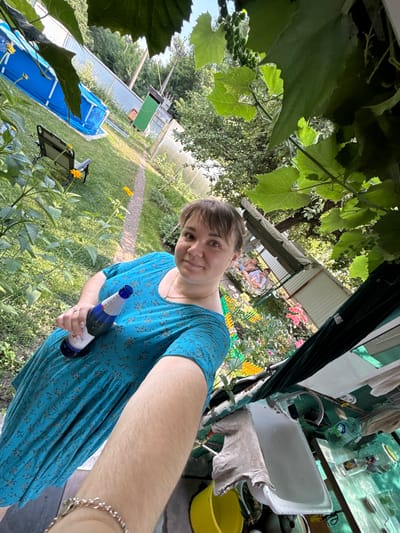 A woman in a blue floral dress holds a bottle of Blanche de Paris champagne while standing in a backyard in Nizhne-Chufichevo, Russia. The scene is set during the day, with sunlight illuminating the lush greenery and a blue inflatable swimming pool in the background. Other objects visible include a lounge chair, a small structure with a green door, and flowering plants. The image appears to be a selfie, with the woman's arm and the bottle of champagne prominent in the foreground.