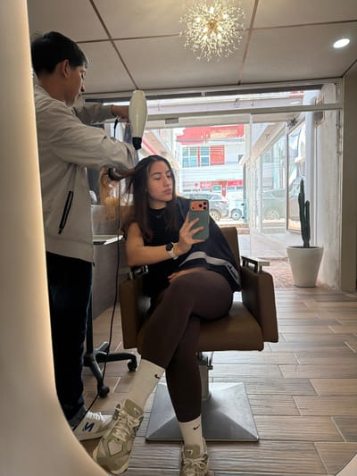 A mirrored reflection captures a woman seated, taking a selfie with her phone. She wears a casual outfit of a t-shirt and leggings. Her legs are crossed and she is sitting in a salon chair. A man stands beside her, holding a hair dryer, seemingly styling her hair. The reflection reveals a bright space with a window showing an outdoor street scene. A chandelier is visible on the left, and a potted cactus sits near the window.