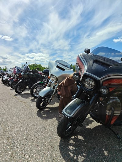 The image depicts a row of motorcycles parked on an asphalt surface, possibly a parking lot or road. The sky is blue with scattered white clouds. The motorcycles are of various colors, including black, silver, and red. A child is standing near one of the motorcycles, partially obscured by the vehicle. The location is possibly near the Borodinskogo Muzeya, Russia, but this cannot be definitively confirmed from the image alone. The scene suggests a motorcycle gathering or event.