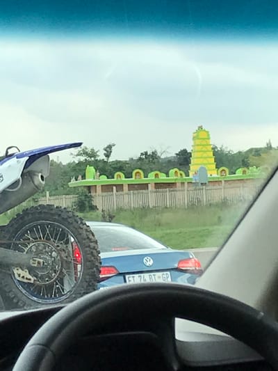 The image displays a roadside view captured from within a vehicle, looking forward through a tinted windshield; a portion of a steering wheel is visible in the lower right. In the foreground, on the left, the rear section of a white and blue motocross dirt bike is present, featuring a knobby tire, spokes, and an exhaust pipe. Ahead in the midground, a blue Volkswagen sedan is visible, bearing the South African license plate "FT 74 RT - GP". In the background, against green hills and an overcast sky, stands a brightly colored Hindu temple, identified as the Shri Radhe Shyam Temple, a landmark visible in the vicinity of Sandton, South Africa. The temple exhibits a prominent, multi-tiered yellow tower (gopuram) with green accents and an extended lower structure in green, adorned with multiple arch-like decorative elements. The scene depicts a road or highway environment.
