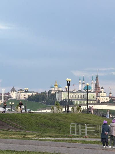 This image captures a section of a city skyline, dominated by a clear blue sky. The buildings, characterized by historical architecture, feature a mix of styles, including onion domes, spires, and classical facades. Among the structures, a prominent green-domed building stands out, while several other buildings exhibit white and beige exteriors with intricate detailing. The scene appears to be a public space, with a paved walkway and a grassy embankment.
Several individuals are present, some walking and others standing. Two people in the foreground, viewed from behind, are wearing jackets and purple headscarves, suggesting a cooler climate or autumn season. One person is holding a phone. Further in the background, other figures are scattered throughout the scene. A white metal fence is visible, separating a grassy area from the walkway. Streetlights with multiple globes are strategically placed along the path.
The lighting suggests it is daytime, likely mid-morning or afternoon, with soft shadows and good visibility. The weather appears to be fair. The presence of lush green grass and trees indicates a temperate climate. The overall atmosphere is one of a peaceful, possibly tourist-oriented, urban environment. The architecture strongly suggests a historical city center in Russia, consistent with the provided location context of Tarlashi, Russia. No visible text is present in the image.