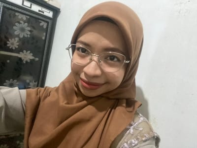 A young woman wearing a brown hijab and glasses is taking a selfie. She is wearing a light-colored top with a textured pattern and a slightly sheer overlay with intricate embroidery. She is smiling and looking towards the camera. The background shows a white wall and a portion of a cabinet with a floral design.