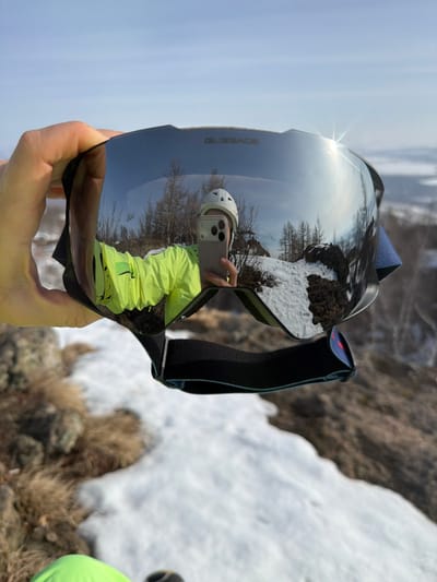 A right hand holds a pair of "GLISSADE" branded ski goggles with dark, reflective lenses, centered in the frame. The goggles' surface reflects a person wearing a white helmet and a bright neon yellow/green jacket, holding a light-colored smartphone to capture a self-portrait. This reflection also displays a clear blue sky, bare deciduous trees, and snow-covered rocky terrain. A prominent sun flare is visible on the upper right portion of the goggle lens. The background directly visible beyond the goggles features a landscape of white snow patches interspersed with dry, light brown grass, exposed rocks, and sparse, bare trees under a pale blue sky. The scene is located outdoors in an elevated, possibly mountainous or hilly environment within Zelenaya Polyana, Russia.