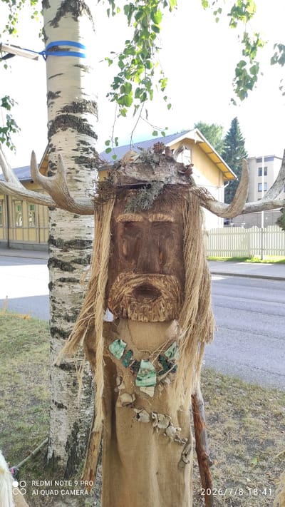 The image shows a close-up of a folk art figure made of wood and natural materials, positioned outdoors in a city setting. The figure is designed to resemble a human face with a beard and long, straw-like hair. It is adorned with large antlers, a headdress made of bark and moss, and a necklace composed of green stones and shells. The figure is dressed in a burlap-like material, with additional natural elements hanging from its body.

Behind the figure, a birch tree is visible, with a blue ribbon tied around its trunk. In the background, a yellow house with a gabled roof can be seen, along with a white picket fence and a paved road. The presence of buildings and a road indicates an urban or suburban environment.

The image also contains a timestamp and text overlay, "REDMI NOTE 9 PRO AI QUAD CAMERA 2026/7/8 18:41". This suggests the photo was taken with a Redmi Note 9 Pro phone on July 8, 2026, at 6:41 PM. While the exact location in Iisalmi, Finland, cannot be precisely determined from the visual information alone, the style of the house and the overall atmosphere are consistent with Finnish scenery. The folk art nature of the figure might suggest it is part of a local festival, exhibition, or a permanent installation in a public space.