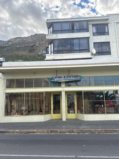 The image captures the exterior of "WAWA Workshop," a surfboard building business, on a street in Muizenberg, South Africa. The building is a multi-story structure with large windows displaying surfboards for sale. The business's sign, shaped like a surfboard, hangs vertically above the entrance. To the left of the building is a mountain under a cloudy sky. A road is visible in the foreground.