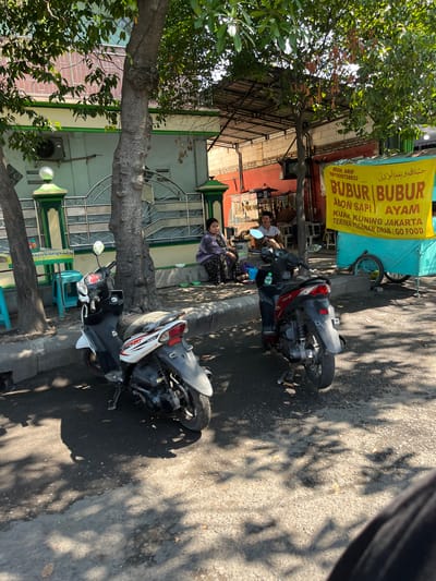In Sumokali, Indonesia, a street scene unfolds under dappled sunlight filtering through trees. Two motorcycles, a white and black one on the left and a red and black one on the right, are parked on the asphalt. Behind them, a woman in a patterned dress and a purple cardigan sits on a bench near a building with a light green facade and decorative metal bars. To the right, a bright yellow banner advertising "BUBUR BUBLR" with Arabic script and Indonesian text hangs from a turquoise food cart. A man in a patterned shirt stands behind the cart, and another person is partially visible. The environment appears to be an urban street with a focus on a food vendor.