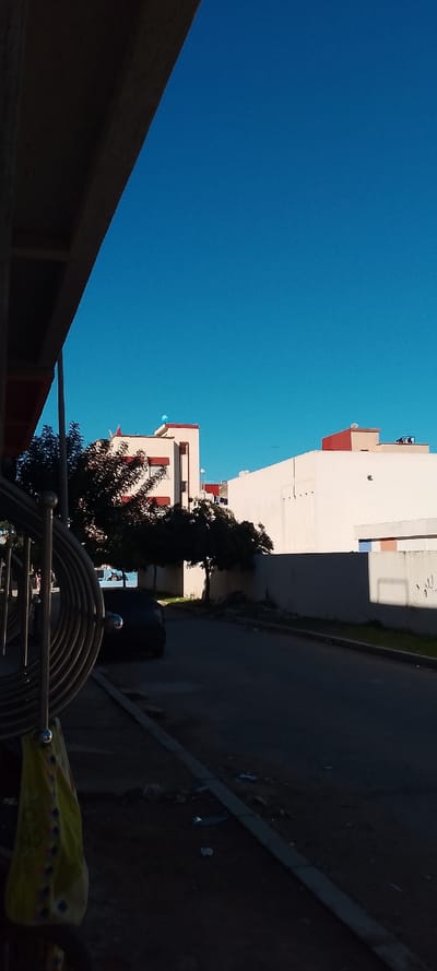 The scene depicts a street in Aïn El Aouda, Morocco, under a clear, bright blue sky. The perspective is from beneath an overhead dark structure, possibly an awning or balcony, visible in the upper left. In the immediate foreground to the left, a silver-colored metal railing or bicycle rack with spiraling elements is prominent, from which a yellow patterned cloth or bag hangs. The ground level comprises a dark, shadowed asphalt road on the right, separated by a concrete curb from a dirt verge on the left. Scattered debris, including papers and plastic, is visible on both the road and verge.
A dark-colored sedan is parked on the left side of the road, partially obscured by deep shadow. Beyond this, several trees with dark, dense foliage line the street. The midground features residential or commercial buildings. The most prominent building, slightly to the left, is light-colored (cream or off-white) with reddish-brown trim around windows and rooflines. A light blue, spherical object, possibly a water tank cover, is visible on its rooftop. Further to the right, another multi-story light-colored building with a high wall exhibits vertical indentations on its facade. Its rooftop features reddish-brown parapet walls and multiple satellite dishes or antennas. Sunlight appears to originate from the upper right, casting significant shadows across the street and foreground elements. No individuals are visible within the frame.