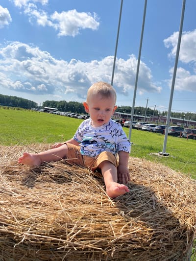 A young child with blond hair and blue eyes is sitting on a large bale of hay. The child is wearing a white t-shirt with a blue and black pattern and brown shorts with a pattern. The child is looking at the camera with a surprised expression. Their left foot is visible, and they are reaching out with their left hand towards the hay.

The setting is outdoors, with a bright blue sky filled with fluffy white clouds. A green grassy field stretches out behind the hay bale, with a line of trees in the distance. Several cars are parked in the field, and there are some buildings visible further back. There are also three vertical poles in the foreground, which are likely part of some sort of structure.

The image appears to have been taken during the daytime, with bright sunlight casting shadows. The overall mood is cheerful and playful, suggesting a fun outdoor activity. There are no visible text elements in the image. The location context provided is Akhlebaikha (Kovriginskii S/S), Russia.