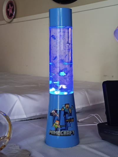 A Minecraft-themed lava lamp with a blue base is standing on a white textured surface. The lamp has a clear cylindrical tube filled with blue liquid and small suspended objects, lit from below by a blue light. The base of the lamp features Minecraft characters and the game's logo. A white cord is plugged into the base of the lamp and extends to the right. To the right of the lamp, a black electronic device, possibly a speaker or charger, is visible. In the background, a white folded sheet or blanket is seen, and a wall with some faint marks is behind it. To the left of the lamp, a portion of a clear glass object with a gold rim is visible. The scene is illuminated by the lamp's blue light and an unseen light source from the right.