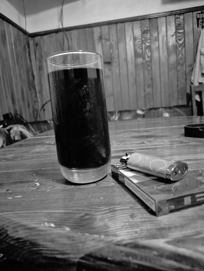 A high-angle, black and white, medium shot captures a glass of dark liquid, condensation beading on its surface, placed on a wooden table. Adjacent to the glass is a pack of cigarettes, with a lighter resting on top. The background features a wooden paneled wall, suggesting an indoor setting, possibly a home or bar. The lighting is dim, casting soft shadows. The scene appears to be set in Iskrets, Bulgaria, as indicated by contextual information not directly visible in the image. There are no people present in the image, and no actions are depicted, beyond the static arrangement of objects. The environment is a still life, evoking a sense of quietude or a moment paused.