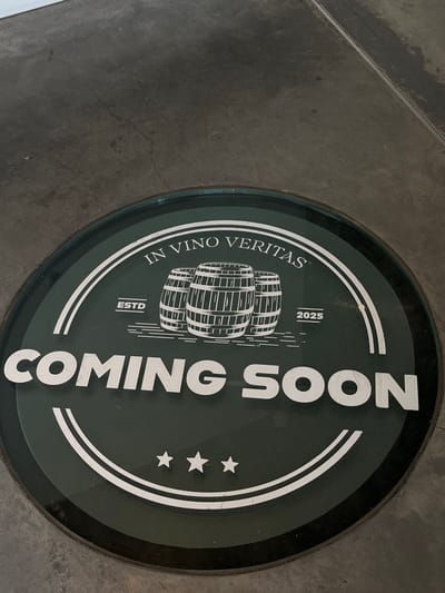 A circular dark green floor decal is embedded in a dark gray concrete surface in Overveen, Netherlands. The decal features white text and graphics. Arced across the top reads "IN VINO VERITAS". Below this, three stylized white wine barrels are depicted horizontally, with "ESTD" to their left and "2025" to their right. Prominently displayed in large white capital letters across the lower center is "COMING SOON". Three small white stars are arranged horizontally near the bottom edge of the decal. The surrounding concrete floor exhibits minor cracks and texture variations. No people, other objects, actions, or interactions are present in the scene.
