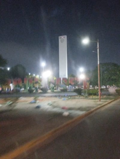 The image is a blurred nighttime shot of a park in Barquisimeto, Venezuela. In the background, a tall, rectangular, white clock tower is visible, illuminated by bright lights, as is a monument that has the word "Barquisimeto" spelled out in large letters. The monument is decorated with colorful paint. Streetlights illuminate the scene. There are a few trees in the park. In the foreground, the edge of a road or curb is visible.