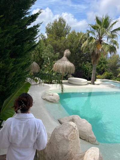 The image depicts a serene outdoor scene in Can Güell, Spain, featuring a person in a white bathrobe from behind, standing near a swimming pool. The pool has clear turquoise water and a sandy, beach-like surround with large naturalistic rock formations. To the left of the frame, a tall, dark green conifer tree dominates the foreground. In the mid-ground, there are thatched umbrellas and lush green vegetation, including palm trees and other tropical plants. A white, circular lounge bed is visible on the sand near the pool. The background reveals a dense forest and a cloudy blue sky. The person's hair is pulled back with a gold clip.