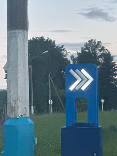 The image shows a blue directional sign with three white chevrons pointing to the right, indicating a path or direction. The sign is mounted on a dark blue base. In the foreground, to the left, is a large, weathered concrete post painted with blue at the bottom. Behind the sign, there is a stand of dark green trees under a cloudy sky. In the middle ground, there are utility poles and a small white circular sign. The ground is covered with green grass. This scene is located in the city of Vereya, Russia.