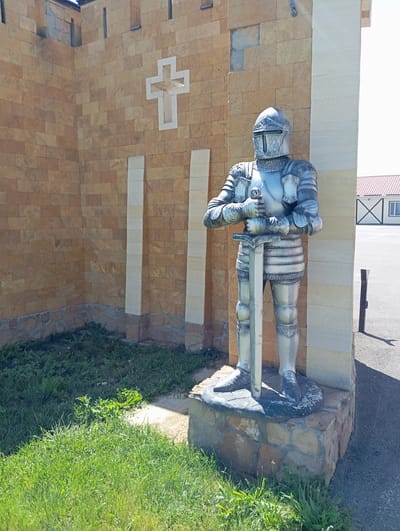 A silver statue of a knight in full armor stands in front of a tan brick wall. The knight is holding a sword vertically in front of him. The wall has a white cross symbol embedded in it. The ground in front of the statue is covered with grass. In the background, there is a building with a red roof and a paved area. The scene is illuminated by sunlight.