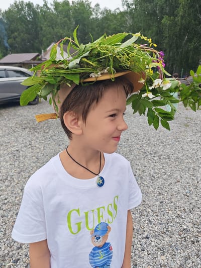 A young boy wearing a white t-shirt with the "Guess" logo and a bear graphic stands outdoors in Mokhovichki, Russia. He has a straw hat adorned with green leaves and white and purple flowers on his head. The boy is looking to his right with a slight smile. The background shows trees, a house with a brown roof, and a car. The ground is covered in gravel.