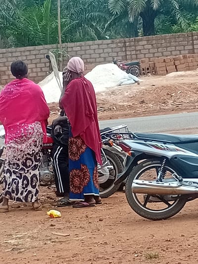 The image captures an outdoor scene in Tuaniako, Nigeria, on a reddish-brown dirt surface adjacent to an asphalt road. Three individuals are standing near motorcycles.
**People:**
1. A female, positioned on the left with her back to the viewer, wears a bright pink top or shawl and a white-and-black patterned skirt. She is standing next to a red motorcycle.
2. A female, centrally located and facing away from the viewer, wears a light pink head covering, a dark red shawl, and a blue skirt with reddish-brown patterns. Her right hand is near her face.
3. An individual, partially obscured between the first two, appears male, wearing dark pants with white vertical stripes. Only the lower torso and legs are distinctly visible.
**Objects:**
* Two motorcycles are prominent in the foreground: a red motorcycle on the left with the text "LONG" visible, and a dark-colored motorcycle on the right with a silver exhaust and black wheel.
* A yellow and orange plastic wrapper lies on the dirt ground in the foreground.
* In the background, beyond a section of grey asphalt road, construction materials are visible. These include a large pile of white aggregate, stacks of red bricks, and a low wall constructed from grey cinder blocks.
* A third, smaller red motorcycle is discernible near the construction materials in the background.
* Dense green foliage, including palm trees, is visible behind the constructed wall.