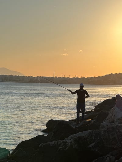 The image depicts a sunset scene in Alimos, Greece. A person is silhouetted against the warm orange and yellow sky, fishing from a rocky shoreline. The individual stands on large dark rocks, holding a fishing rod extended over the calm sea. The water reflects the golden light of the setting sun. In the background, the cityscape of Alimos is visible as a dark silhouette against the horizon, with buildings, some masts of sailboats, and industrial structures like chimneys and cranes. A mountain range can be seen in the far distance to the left. The sky has a gradient of color, transitioning from a lighter yellow at the top to a pale blue towards the horizon, with a few wispy clouds. A thin contrail from an aircraft is visible in the upper portion of the sky. Another person is partially visible to the right, also on the rocks, facing away from the camera. The overall atmosphere is tranquil and serene, characteristic of a coastal sunset.