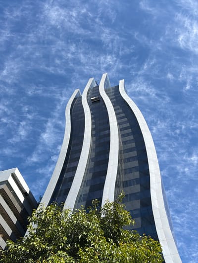 A low-angle shot captures a modern, curved architectural building against a bright blue sky with wispy white clouds. The building features a dark, glass-paneled facade interspersed with white, wave-like horizontal bands. A lush green tree with dense foliage partially obstructs the view of the building on the right side. In the lower right corner, a portion of another building is visible, with a grey and white geometric design. The lighting suggests it is daytime, likely with sunny weather. There is no visible text in the image. The scene depicts an urban environment, possibly in Asuncion, Paraguay, characterized by contemporary architecture and natural elements.