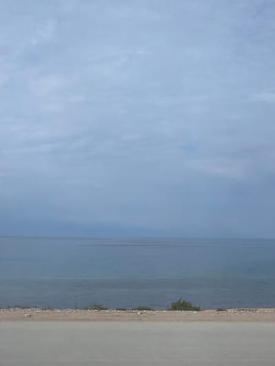 This image depicts a serene seascape under a cloudy sky. The horizon line is clearly defined, separating the vast expanse of the ocean from the overcast sky. The water appears calm, with subtle ripples indicating a gentle breeze. The foreground features a narrow strip of land, possibly a beach or embankment, with sparse vegetation. A road or pathway runs along the very bottom of the frame. The color palette is dominated by muted blues and grays, contributing to the tranquil atmosphere. There are no people or specific structures visible in this image. The location is not in the city of Tong, Kyrgyzstan, as this appears to be a coastal scene.