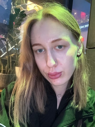 A woman is captured in a close-up shot, slightly tilted. Her hair is blonde with hints of green. The woman has fair skin and blue eyes, with a small stud piercing on her nose. A small heart tattoo is visible on her neck. She is wearing a black shirt or top underneath a green jacket.
The background includes a blurred screen or display with text in a Cyrillic alphabet. There is also vegetation visible, likely plants, and architectural elements that suggest an indoor space, possibly a restaurant or public venue. The image was taken in Ol'ginskaya, Russia.