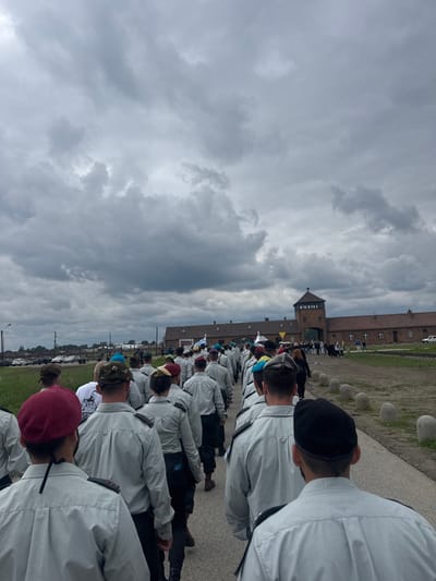 The image shows a group of people walking in a line towards the main gate of the Auschwitz-Birkenau Memorial and Museum in Brzezinka, Poland. The location is a former Nazi concentration camp during World War II. The people are dressed in gray shirts and pants, with some wearing hats. They are walking on a paved path, with green grass and trees on either side. In the background, there is a brick building with a tall tower, and there are other people walking in front of the building. The sky is overcast with gray clouds. The image suggests a solemn and somber atmosphere, likely capturing a moment of remembrance and reflection.