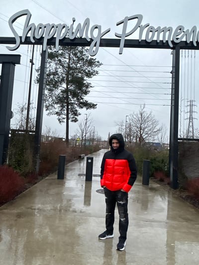A person stands on a wet pavement under a metal sign that reads "Shopping Promenade." The person wears a black hooded jacket with an orange puffy vest. A few bare trees are visible in the background along with telephone poles. The sky is overcast, adding to the dreary atmosphere.