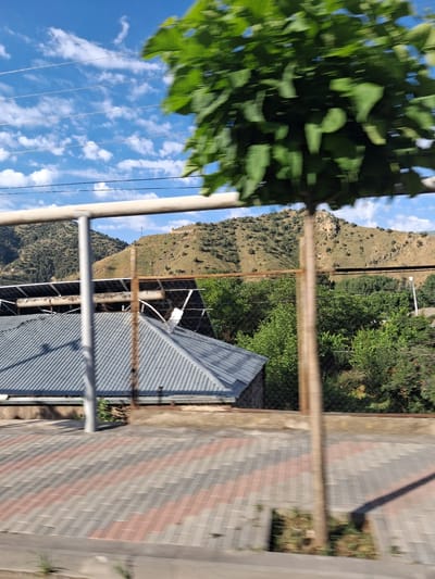 The image depicts a view from a moving vehicle on a sunny day in Vayk, Armenia. In the foreground, a paved area with a pattern of grey and reddish-brown rectangular pavers is visible. A thin, wooden pole, likely a fence post, is positioned slightly to the right of the frame's center. Behind the pole and pavers, a grey, corrugated metal roof of a building is seen, extending to the left. Above the roof, a cluster of solar panels is mounted. A chain-link fence separates the building from the background.

Further in the background, a dense line of green trees forms a boundary. Beyond the trees, a treeless, undulating hillside is covered in sparse vegetation and scattered rocks. The hillside appears to be a natural landscape, possibly a mountain. The sky above is a bright blue, dotted with scattered white clouds. Several power lines are strung horizontally across the sky. A blurred green tree with broad leaves occupies the upper right portion of the image, suggesting it is close to the viewpoint and partially obstructing the view. The overall scene conveys a rural or semi-rural environment within Vayk.
