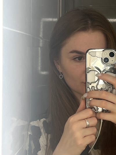 A young woman with brown hair and light skin takes a selfie in a bathroom with tiled walls. She is holding a silver phone with a skull-patterned case up to her face, covering the right side of her face. She is wearing a silver ring on her left ring finger and a small hoop earring in her left ear. The background shows a reflective surface that appears to be a mirror or a metallic wall. There is also a white cord visible near her hand, possibly from headphones. The location is not specified in the image, so it cannot be determined to be in Нижние Муллы, Russia.