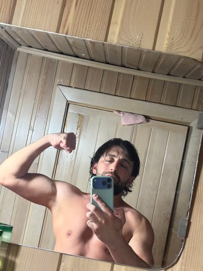 A bare-chested man is posing in front of a mirror while holding a smartphone. The reflection shows his face and flexed arm. He appears to be in a room with wood paneling. The location in the city of Тартат, Russia, is not discernible from the image.