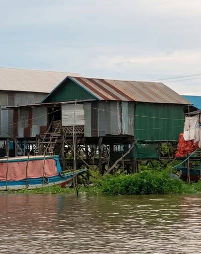 This is a medium shot, eye-level, outdoor, daytime, vertical photograph of a stilted village built over water. The structures are constructed primarily from corrugated metal and wood. In the foreground, the murky water of a river or lake occupies the lower half of the frame, reflecting the overcast sky. A portion of a blue and red boat with a pink canopy is moored to the left. Behind the boat, a complex of stilted houses is visible. One prominent house in the center features a green corrugated metal facade and a rusted corrugated metal roof with a brown patina. A wooden staircase ascends to the entrance of this structure. To the left of this house, another similar structure is partially obscured. Dense green vegetation, likely aquatic plants, grows around the base of the stilted buildings. The sky is cloudy and pale. No people are visible in the scene. The location is identified as the village of Soutr Nikom in Cambodia.