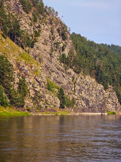 A wide river flows in the foreground of the image. The river has a choppy surface with reflections of the surrounding environment. On the right bank of the river, a steep, rocky cliff rises abruptly. The cliff face is rough and textured, with various shades of gray and brown rock. Patches of green vegetation, including trees and shrubs, cling to the rocky slopes and the top of the cliff. Pine trees, in particular, are visible scattered across the cliff face and densely covering the area behind it. The sky above is a clear, pale blue with a few faint wisps of clouds. The overall scene depicts a natural landscape characterized by the juxtaposition of the flowing water and the rugged, vegetated cliff. This location is in Beregovaya-Podemnaya, Russia. There are no people or man-made structures visible in the image.