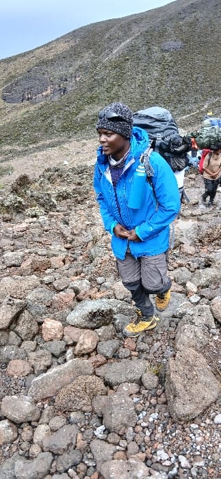 The image displays a high-altitude scene on a rocky trail at Gilman's Point, Tanzania, under an overcast sky. In the foreground-midground, an individual with dark skin walks uphill, oriented slightly right. This person wears a dark knitted beanie with sunglasses perched on top, a bright blue hooded jacket over a darker inner layer, grey cargo-style pants with black lower leg sections, and yellow hiking boots. A light grey backpack is on their back, and a blue water bottle is clipped to the left shoulder strap. Their hands are clasped at waist level. Further up the trail to the right, two additional individuals are visible, also ascending, each carrying large, bulky backpacks; one pack is green/camouflaged, and another is dark. The ground consists of uneven dark, grey, and reddish-brown rocks and scree, with sparse, low-lying vegetation interspersed. A barren, vegetated mountain slope forms the background under a light grey sky.