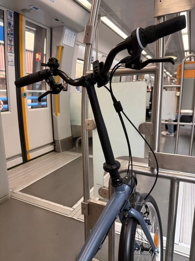 A light blue/grey folding bicycle is depicted upright within the clean, modern interior of a public transport vehicle, likely a train or tram, in Pasaia, Spain. The image is captured from a low angle, focusing on the bicycle's handlebars and front wheel.

The bike's black handlebars feature textured black grips with white rings, along with black brake levers and a shifter on the left side. Black cables run neatly from the controls, along the bicycle's frame. The frame itself is a light blue-grey color, with the brand "BTWIN" clearly visible. Part of the front wheel, including a black tire, spokes, and a reflective orange element, is visible towards the right of the frame.

The interior of the vehicle is characterized by polished metal handrails and supports, white wall panels, and a grey floor. Reflections from overhead lighting are visible on the metal surfaces and a clear panel that may serve as a partition. In the background, a seated person's legs, wearing white sneakers, are partially visible. Below the white panels, there are faint reflections in what appears to be a window or another panel. Some indistinct signs and pictograms are visible on the walls, but the text is unreadable. The scene conveys the practical activity of transporting a bicycle on public transport.