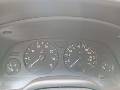 This is a close-up, eye-level shot of a car's dashboard in Bugulma, Russia. The focus is on the instrument cluster, which is dimly lit and appears to be viewed from the driver's seat. The dashboard itself is a dark, matte material, likely plastic, typical of a vehicle's interior.
The primary objects in the image are the gauges. On the left, a temperature gauge reads slightly below 80 degrees Celsius, indicated by a needle pointing towards the left side of the dial. To the right of this is the tachometer, with a needle pointing just above the "0" mark, indicating the engine is idling. The tachometer is marked in units of "x100 min⁻¹".
The central and largest gauge is the speedometer, with a needle indicating a speed of approximately 20 km/h. The speedometer is marked in increments of 20 km/h, up to 220 km/h. To the right of the speedometer is the fuel gauge, with a needle indicating approximately half a tank of fuel, marked as "1/2".
There are no people visible in the image. The setting is clearly the interior of a car. The lighting suggests it could be daytime, but the interior is somewhat dark, possibly due to interior lighting being on or overcast conditions outside. There are no specific weather cues or strong emotional indicators. The visible text consists of the numbers and units on the gauges: "80", "100", "°C", "30", "40", "50", "60", "70", "20", "x100", "min⁻¹", "80", "100", "120", "140", "160", "180", "200", "220", "km/h", "km", "1/2", and "1/1". The general appearance of the dashboard suggests it might be from an older model car.
