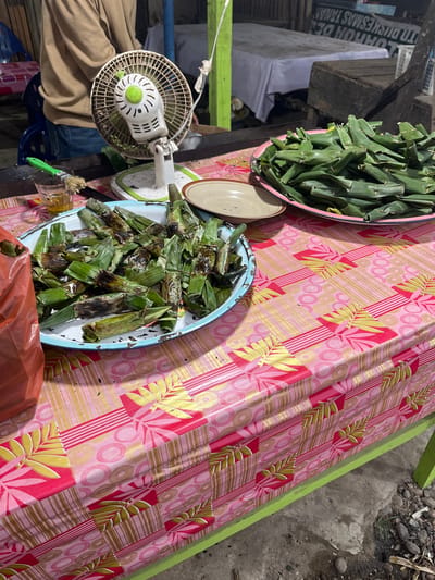 An outdoor food stall in Peurupok, Indonesia, displays grilled snacks wrapped in banana leaves. The table is covered with a vibrant pink and yellow floral-patterned tablecloth. In the foreground, a blue-rimmed metal tray overflows with these grilled parcels, some showing signs of charring from the grill. Next to it, a large pink plastic bag sits beside a small glass containing a yellowish liquid and a green brush with bristles. A white electric fan, with a green accent on its controls, stands behind the tray. Another plate, piled high with uncooked, similarly wrapped snacks, is positioned to the right of the fan. In the background, a white-clothed table and wooden crates are visible, hinting at the market setting. A person, partially visible from the chest down, is wearing a tan long-sleeved shirt and blue jeans, suggesting they are the vendor. The ground beneath the stall is a mix of concrete and gravel.