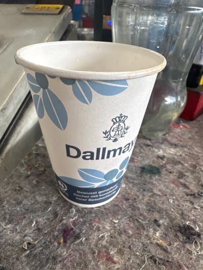 This media file captures a close-up, rotated view of a disposable paper coffee cup with a blue floral design. The cup appears to be resting on a textured, grey surface, possibly a felt or fabric material, with scattered colorful fibers. Behind the cup, blurry elements suggest a background setting, including what looks like a glass bottle and a red object.
The cup itself is white with light blue floral patterns. Prominently displayed on the cup is the "Dallmayr" logo, a circular emblem with a crown and intricate detailing, followed by the brand name in a dark, sans-serif font. Below the brand name, there is text in German: "Bewusst genießen Becher mit kur freier Beschr," which translates to "Consciously enjoy cup with short free besch." The "E" in the Dallmayr logo is also visible.
The overall lighting suggests an indoor environment. The angle of the photograph and the focus on the cup imply that the cup is the primary subject of interest, with the background providing context. No people are visible in the frame. The scene is static, with no discernible activity taking place. The location context provided is Ellwangen, Germany, though no specific visual cues within the image directly confirm this.
