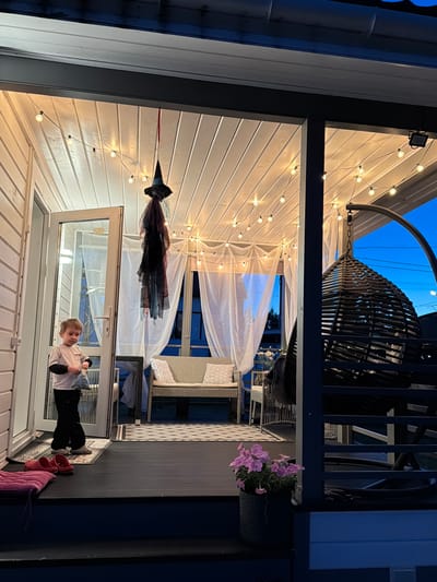 In the city of Alyoshkino DP, Russia, on a porch at dusk, a young boy stands near a glass door, holding a bag. He is wearing a light-colored long-sleeved shirt and dark pants. The porch is decorated with string lights along the white wooden ceiling and a hanging witch decoration. A wicker hanging chair is visible to the right, and a bench with cushions sits further back. A potted plant with pink flowers is placed on a lower level of the porch. The scene appears to be set for Halloween or a similar festive occasion.