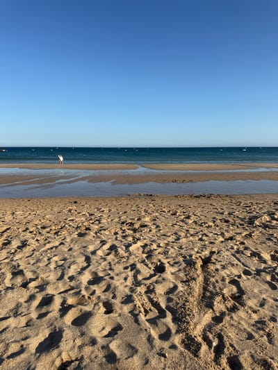 The image depicts a beach scene in Lagos, Portugal, likely taken during daylight. The foreground is dominated by sandy beach with many footprints and a visible sand track. Beyond the sand, a shallow body of water reflects the clear blue sky, separating the beach from the deeper ocean. In the distance, a person wearing a white shirt and red shorts stands in the shallow water, facing away from the camera. The ocean stretches to the horizon, marked by a thin line of land or distant boats and buoys. The overall atmosphere is serene and calm.