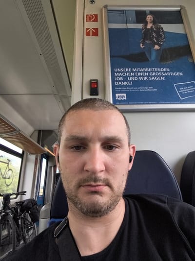 The image depicts a man, likely in his late 20s or early 30s, seated inside a train carriage. He has a shaved head and a short beard, and is wearing black earbuds and a black t-shirt. The man is looking directly at the camera, and his upper torso and face are in the foreground. Behind him, on the right side of the image, is a poster with text in German that appears to be an advertisement or announcement. On the left side of the train carriage, two bicycles are visible, one parked upright and the other leaning against a wall. The environment suggests a public transportation setting, likely a train, with windows revealing a blurred outdoor scene. The poster and bicycles suggest that the train may accommodate passengers traveling with bicycles. The text on the poster, "UBB Usedomer Bäderbahn GmbH" indicates the train operator, which serves the Usedom island region of Germany. Züssow is a municipality in Mecklenburg-Vorpommern, a state in northeastern Germany, and it is plausible that this train is operating within or near this region, given the information on the poster.