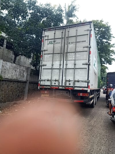 A truck with "J&T CARGO" on its back is stopped on a road with a motorcycle to its right. The truck's doors are white with some graffiti and the phone number 021-0661666. On the left, there is a stone wall with trees looming overhead. The ground is dry and slightly uneven. The license plate number is B 9410 UZB.