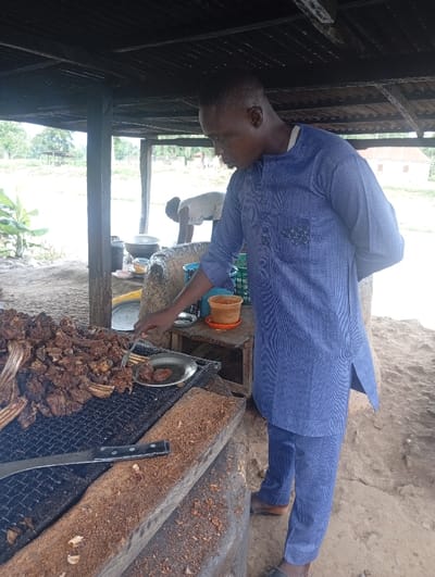 The image displays an outdoor grilling and food preparation area under a rudimentary shelter in Assai Sabongida, Nigeria. A dark-skinned male, estimated to be between 20 and 40 years old, wearing a dark blue patterned tunic and matching trousers, stands on the right side of the frame. He is actively transferring a piece of grilled meat from a large black metal grill onto a small silver plate using a metal fork held in his right hand. To the left on the grill, a substantial quantity of dark brown grilled meat, primarily ribs, is present. A large knife with a black handle rests on a wooden surface beneath the grill, which is covered in light brown particulate matter, likely wood shavings or char. Behind the primary subject, a small wooden table holds a brown clay pot, a red plastic container, and additional metal dishes. In the background, another individual, partially obscured and bent over, is visible near a large metal pot. The ground is exposed, dry, light brown earth. The shelter is constructed with visible wooden posts supporting a dark, planked roof. Beyond the immediate structure, an open, vegetated field extends, with a small building featuring a reddish-brown roof discernible in the distance to the right.
