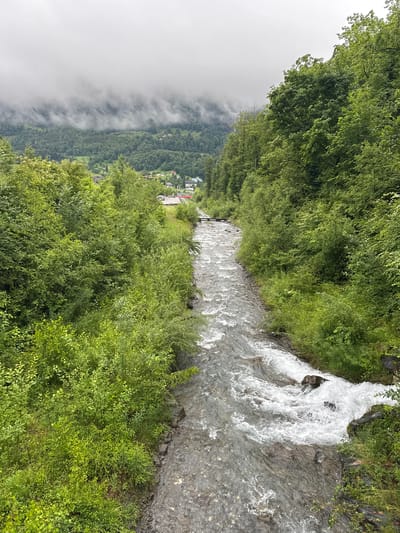 The image shows a fast-flowing river with rocky banks and lush green vegetation on either side. In the background, there are rolling hills covered in trees, with a village nestled in the valley. The sky is overcast, with mist or clouds obscuring the distant mountain peaks. The scene appears to be in a rural or mountainous area, possibly the Swiss Alps, given the mention of Hüsen, Switzerland. There are no people visible in the image. The river flows from the background towards the foreground, with whitewater rapids indicating its swift current.