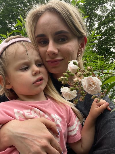 In a close-up, outdoor selfie taken from a slightly low angle, a woman with blonde hair and subtle makeup embraces a young child. The woman's head is tilted down, and she holds a small sprig of light pink roses with buds. The child, with blonde hair and a pink headband, leans into the woman's embrace, looking slightly away from the camera with a neutral expression. The child wears a pink t-shirt with white stripes and white text that appears to say "READY". The woman wears a dark-colored top, and a hint of another person's arm is visible on the right. The background consists of lush green foliage and trees, suggesting a garden or park setting. The lighting is natural and diffused, indicative of daytime. The image captures a moment of tenderness and connection between the two individuals amidst a natural environment. There are no explicit indicators of the city of Knevichi, Russia within the image itself.
