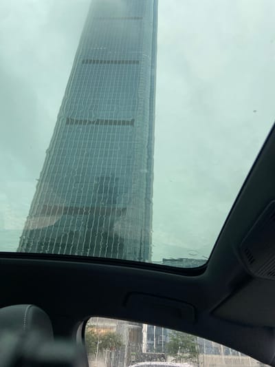 An exterior, eye-level, full shot shows a tall, modern skyscraper through the sunroof of a vehicle. The skyscraper, which has a reflective, glass facade, appears to be distorted by water droplets or streaks on the glass. The building is predominantly gray and blue, with visible horizontal and vertical lines indicating its structure. The sky is overcast and a pale gray. The interior of the vehicle is visible in the foreground, with a black leather seat and parts of the car's roof lining. The scene appears to be in a city, possibly Luohu District, China, given the architectural style of the skyscraper and the overall urban environment suggested by reflections in the building's facade.